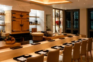 An elegant, modern omakase sushi bar interior featuring a minimalist light-wood L-shaped dining counter lined with cushioned tan chairs, neatly set with black tray placemats, white cloth napkins, and serving boards in front of a chef's preparation area with a traditional wooden icebox.