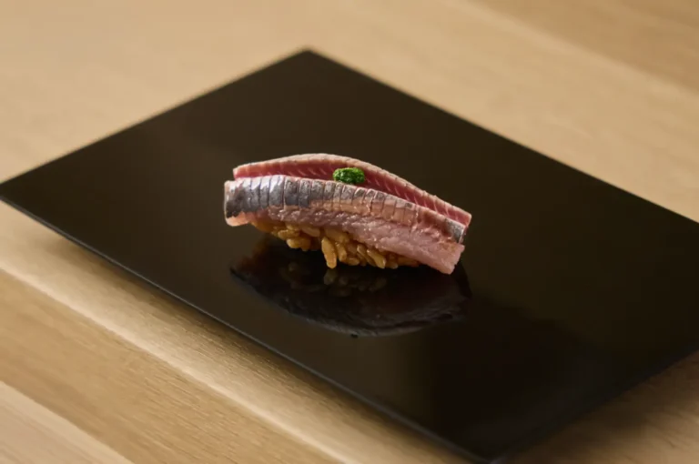 A piece of nigiri sushi featuring meticulously scored silver-skinned fish garnished with a dab of green paste sits atop seasoned brown rice on a sleek black rectangular plate, set against a light wood counter background.