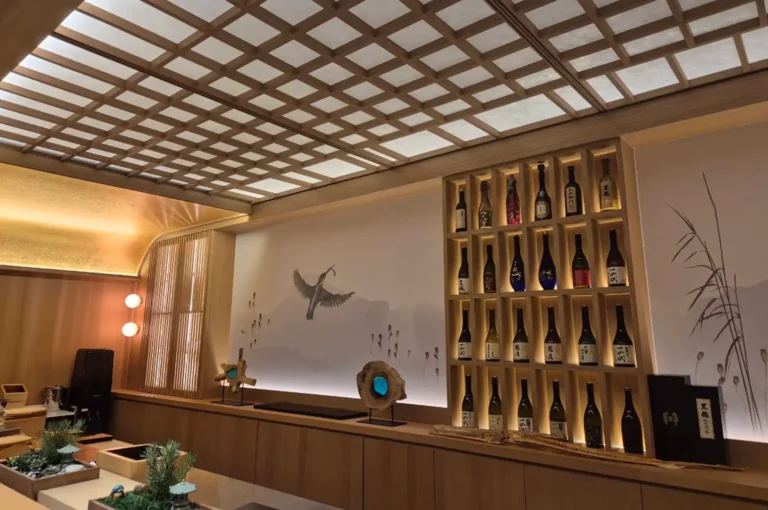 The warm, elegant interior of a Japanese dining room showcases light wood paneling, a grid-patterned illuminated ceiling, and a featured wall mural depicting a flying kingfisher. Built-in wooden shelving displays a curated collection of sake bottles illuminated by warm lighting above a minimalist service counter decorated with miniature potted plants.