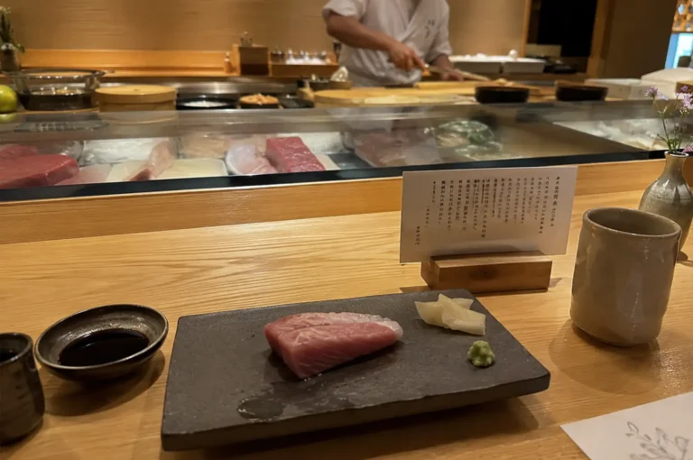 A warm, candid view from a seated diner’s perspective at a wooden omakase sushi counter. In the foreground rests a dark, rustic rectangular plate holding a thick slice of raw, marblized pink fish sashimi, pickled ginger, and a mound of fresh wasabi, flanked by a small bowl of soy sauce and a neutral ceramic teacup. Behind the serving plate, a printed menu card in a wooden holder sits in front of a glass display case showcasing fresh primal cuts of seafood, while a sushi chef wearing a white uniform and face mask works in soft focus in the background.