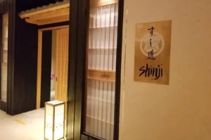 The entrance of a traditional Japanese restaurant features light-toned wooden sliding doors framed by dark wood paneling, subtly illuminated from within and by a warm floor lantern. To the right, a textured cream wall displays a golden brass plaque featuring Japanese calligraphy alongside the restaurant name "Shinji" rendered in a black, hand-drawn script.