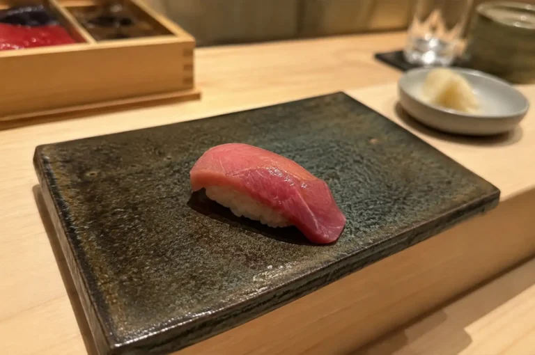 A close-up, eye-level perspective shot captures a single piece of nigiri sushi featuring a glossy, pink-and-red slice of tuna brushed with a light glaze over seasoned rice. The sushi rests centered on a dark, textured rectangular ceramic serving plate on a light wood omakase counter, with a small dish of pickled ginger, a water glass, and a green teacup softly blurred in the background.