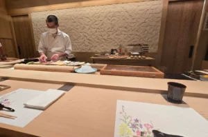 A Japanese omakase chef wearing a white uniform and face mask carefully prepares fresh fish behind a light wooden sushi counter, set against a carved geometric wooden wall in a modern, serene dining room setup.