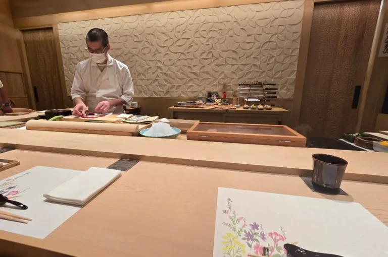 A Japanese omakase chef wearing a white uniform and face mask carefully prepares fresh fish behind a light wooden sushi counter, set against a carved geometric wooden wall in a modern, serene dining room setup.