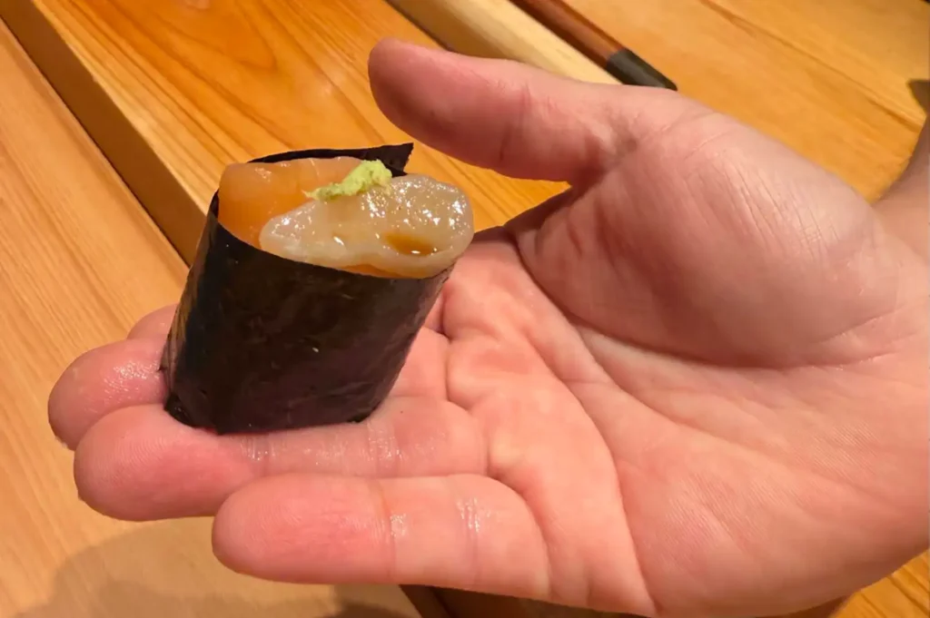 A close-up shot showing a piece of gunkan maki sushi held delicately in the palm of an open hand against a smooth wooden counter background. The piece is wrapped in a dark sheet of nori seaweed, filled with seasoned sushi rice, and topped with delicate slices of fresh seafood dabbed with a speck of green wasabi and soy glaze.