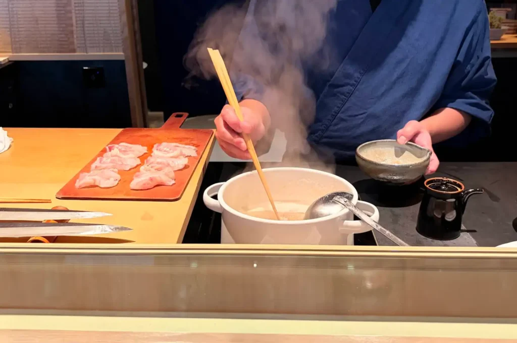 A behind-the-counter action shot depicts a sushi chef clad in a blue traditional uniform using long wooden chopsticks to stir broth steaming inside a white pot on an induction burner. To the left, a wooden cutting board holds carefully arranged slices of raw white fish alongside chef knives, while a small ceramic bowl sits nearby for preparation.