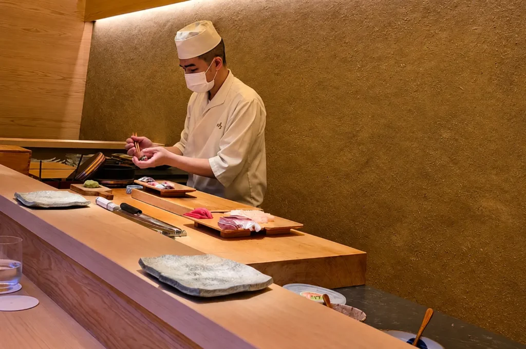 A wide shot of a Japanese sushi chef wearing a white traditional uniform, chef cap, and face mask behind a light wooden omakase counter, meticulously assembling nigiri with chopstick-like culinary tweezers in front of a textured, warm ochre wall.
