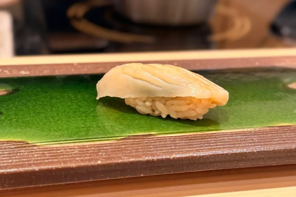 A close-up shot of a piece of delicate white fish nigiri sushi with light-colored seasoned rice, artfully presented on a rectangular dark ceramic plate with a glossy green glazed center.