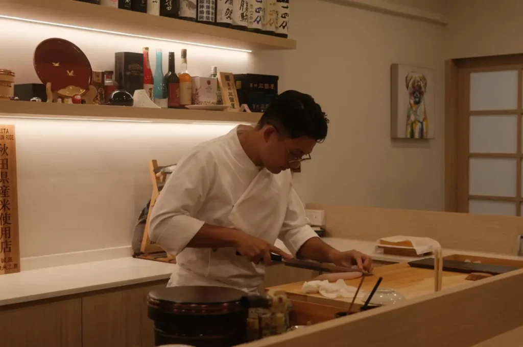 A sushi chef dressed in a white uniform coat stands behind a light wood omakase counter, intently using a yanagiba knife to slice fresh fish, with warm ambient backlighting, sake bottles on shelves, and a wooden rice tub nearby.