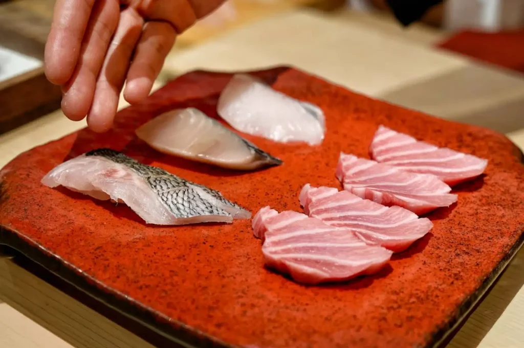 A chef's hand presents an array of fresh, high-grade sashimi arranged on a textured red ceramic platter, highlighting delicate cuts of white fish, silver-skinned fish, and heavily marbled pink tuna slices ready for preparation.