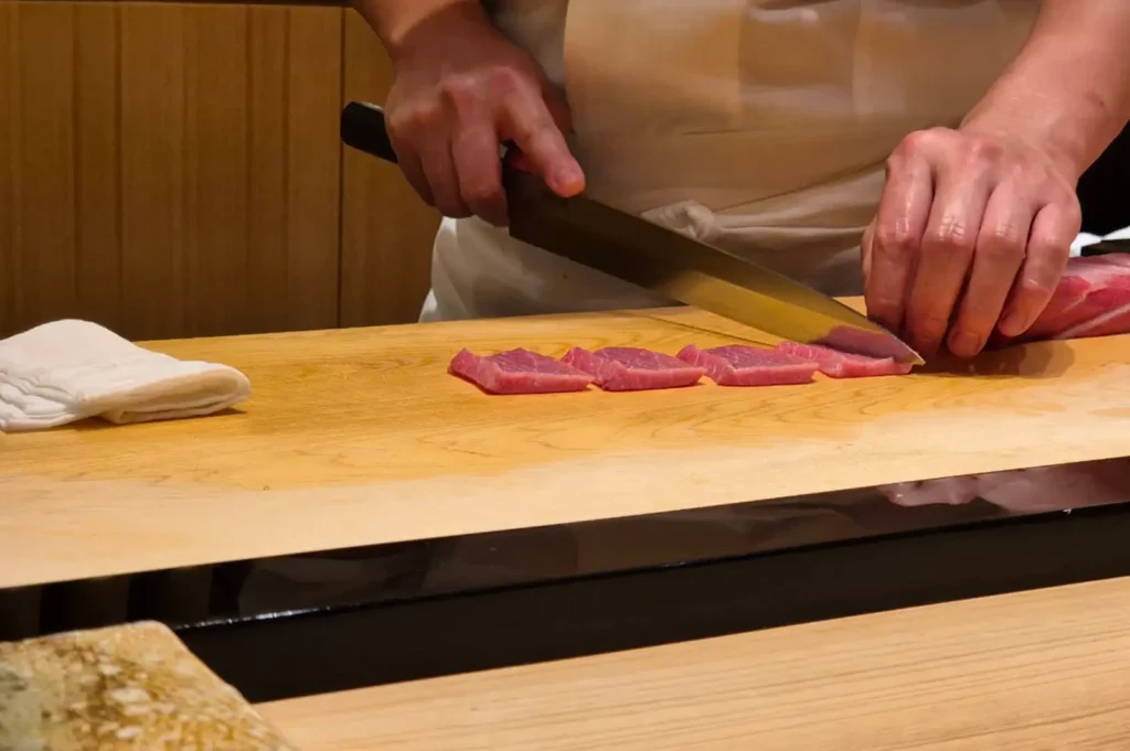A close-up shot captures a chef in a white uniform using a sleek yanagiba sushi knife on a pristine wooden cutting board to slice rich, marbled pink tuna. Four freshly cut pieces of otoro are neatly aligned in a row along the counter, beside a folded white cotton towel.