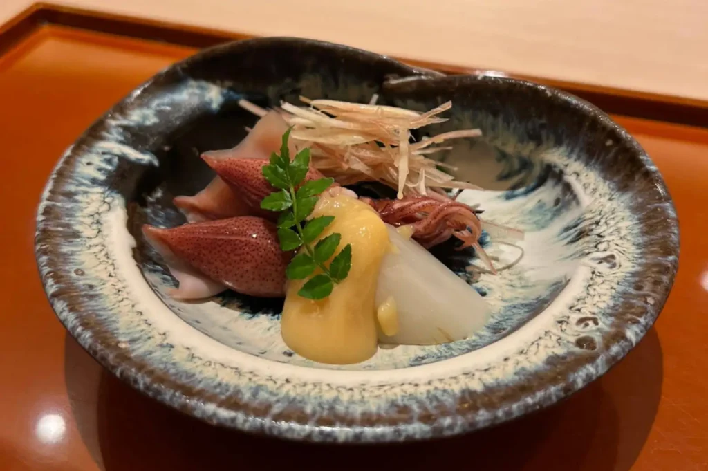 A rustic ceramic bowl with a blue-and-white glazed interior holding tender firefly squid, a slice of white radish, a creamy yellow sauce, thinly shredded ginger, and a small green shiso leaf garnish.