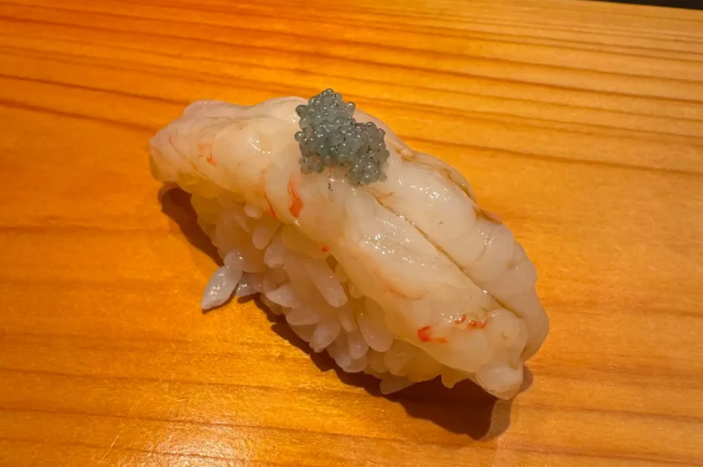 A detailed close-up shot of a single piece of sweet botanical shrimp (amaebi) nigiri resting directly on a clean, light-grained wooden sushi counter. The sweet, translucent white-and-pink shrimp curves over the hand-pressed sushi rice ball and is topped with a small cluster of bluish-grey caviar as a garnish.