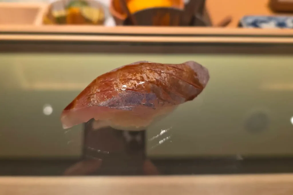 A close-up shot showcases a piece of shiny nigiri sushi served on a glossy black display platter inside a dimly lit omakase setting. The slice of fish exhibits a iridescent sheen with fine score lines, glistening with a light coat of sauce over pressed sushi rice.