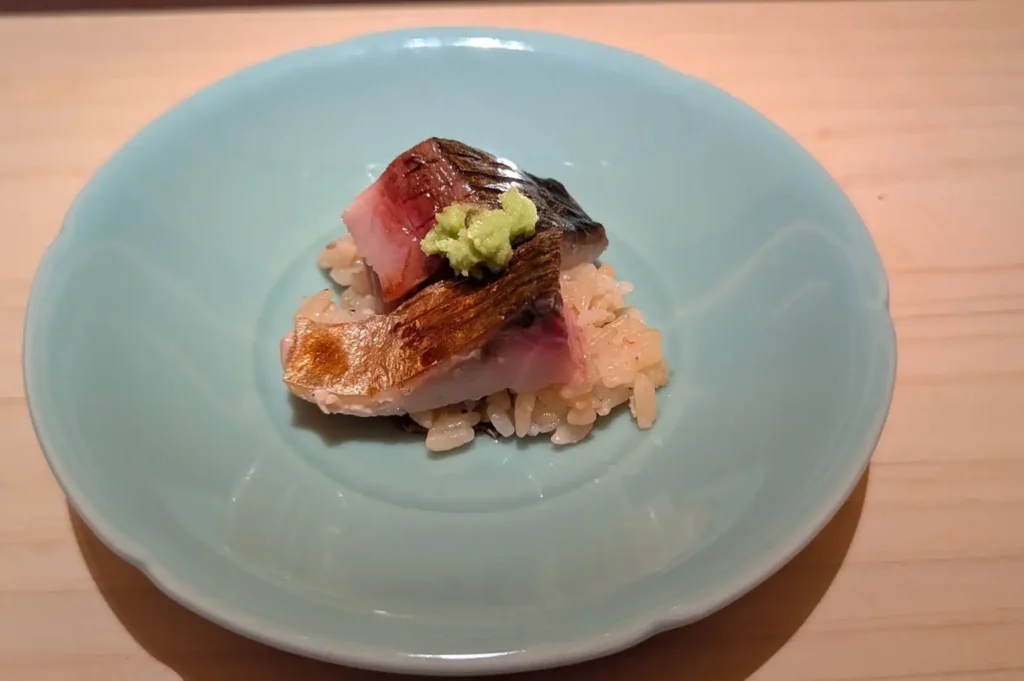 A top-down shot showing a piece of lightly seared, glistening mackerel skin served over a small mound of seasoned sushi rice in a shallow, soft robin’s-egg blue ceramic dish, topped with a dollop of freshly grated green wasabi.