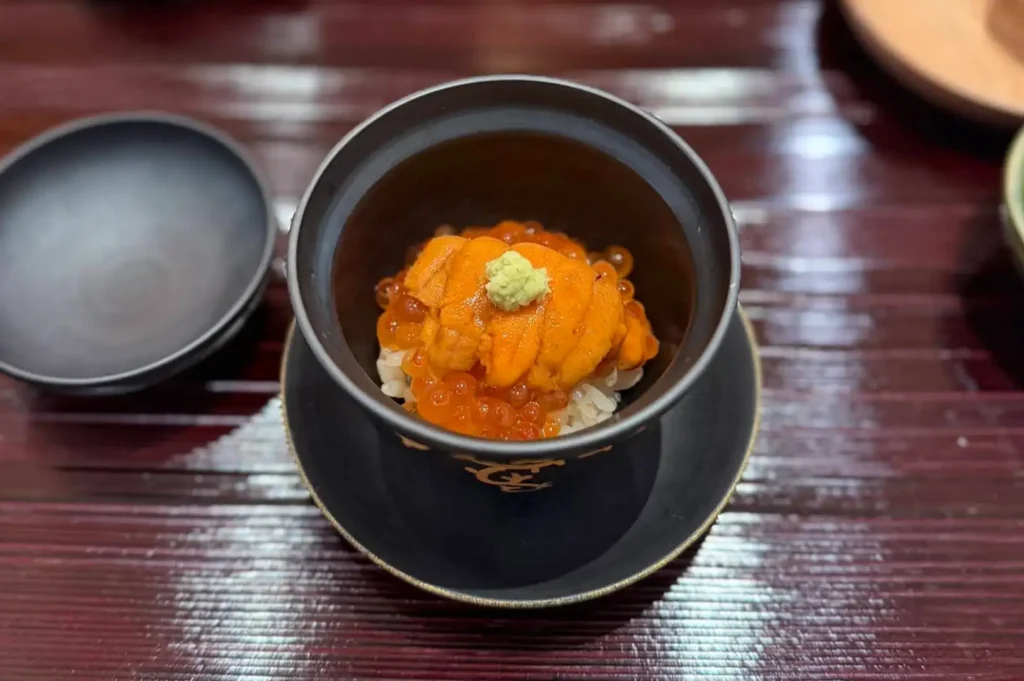 An overhead view of a mini Donburi rice bowl served in a black ceramic dish with gold accents, generously topped with bright orange sea urchin (uni), glistening salmon roe (ikura), and a small dollop of fresh wasabi.