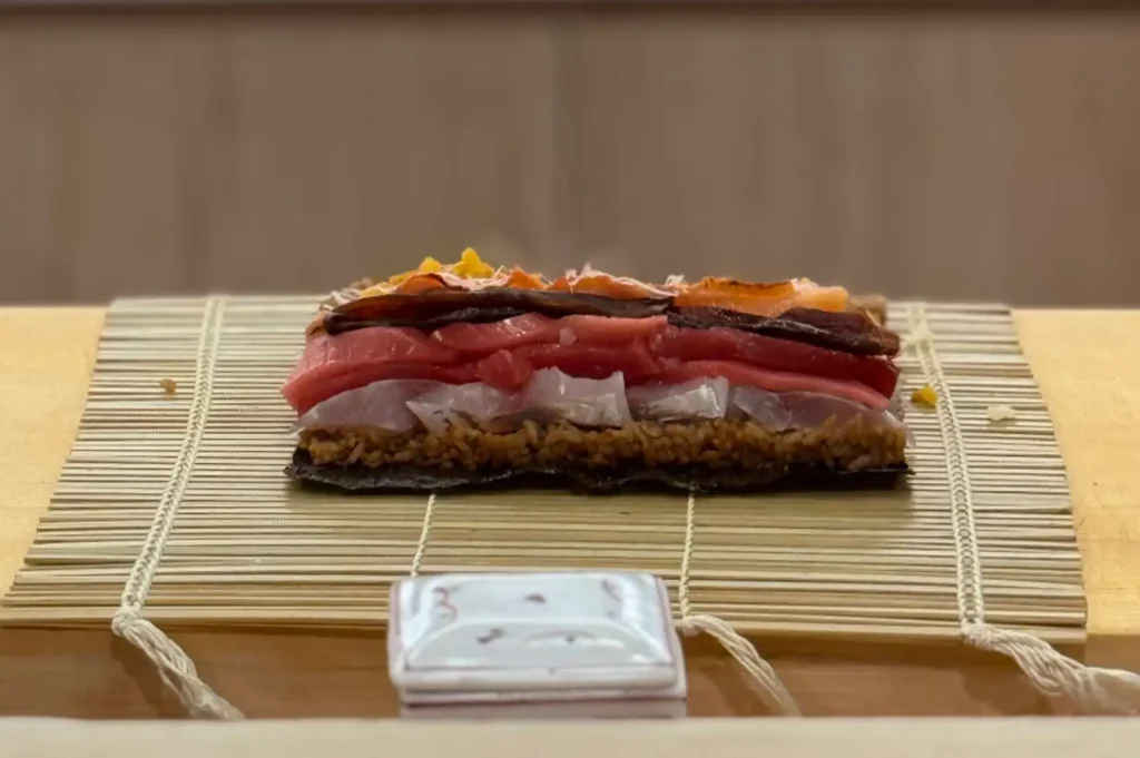 A side view reveals the cross-section of an unrolled sushi roll assembly resting on a bamboo rolling mat, showcasing layered ingredients over dark seaweed and seasoned brown shari, including white fish, vibrant red tuna, dark seared fish, and yellow pickled toppings.