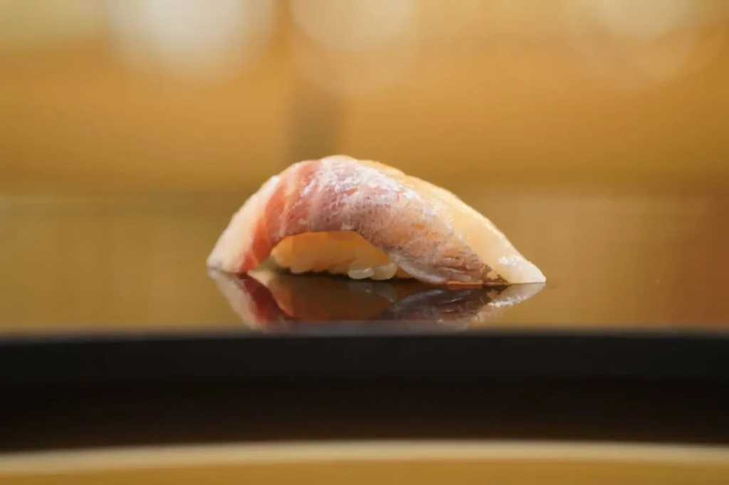A single piece of aged nigiri sushi rests on a high-gloss black tray, casting a soft reflection on the smooth surface. The glistening slice of fish features a delicate gradient of pale pink to silver-streaked red, draped precisely over a hand-pressed mound of seasoned sushi rice against a warm, softly blurred background.