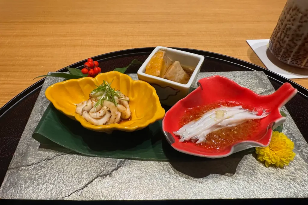 An artistic Japanese appetizer course is presented on a textured silver plate set inside a round black tray, featuring three distinct ceramic dishes. A yellow leaf-shaped bowl holds seared white fish with shredded scallions, a white square bowl contains simmered ingredients, and a red fish-shaped dish displays delicate whitebait resting in a jellied sauce, garnished with red winter berries and a yellow flower.