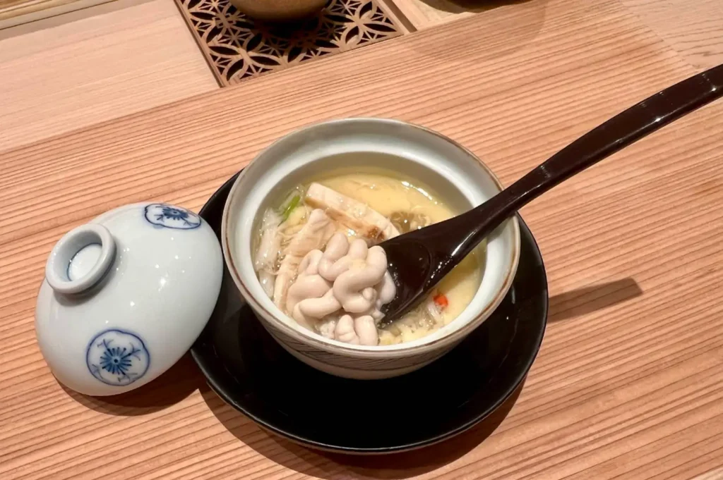 A delicate blue-and-white porcelain cup containing steaming hot chawanmushi topped with cod milt (shirako) and crab meat, accompanied by a dark wooden spoon resting inside and the cup lid set to the side on a light wood counter.