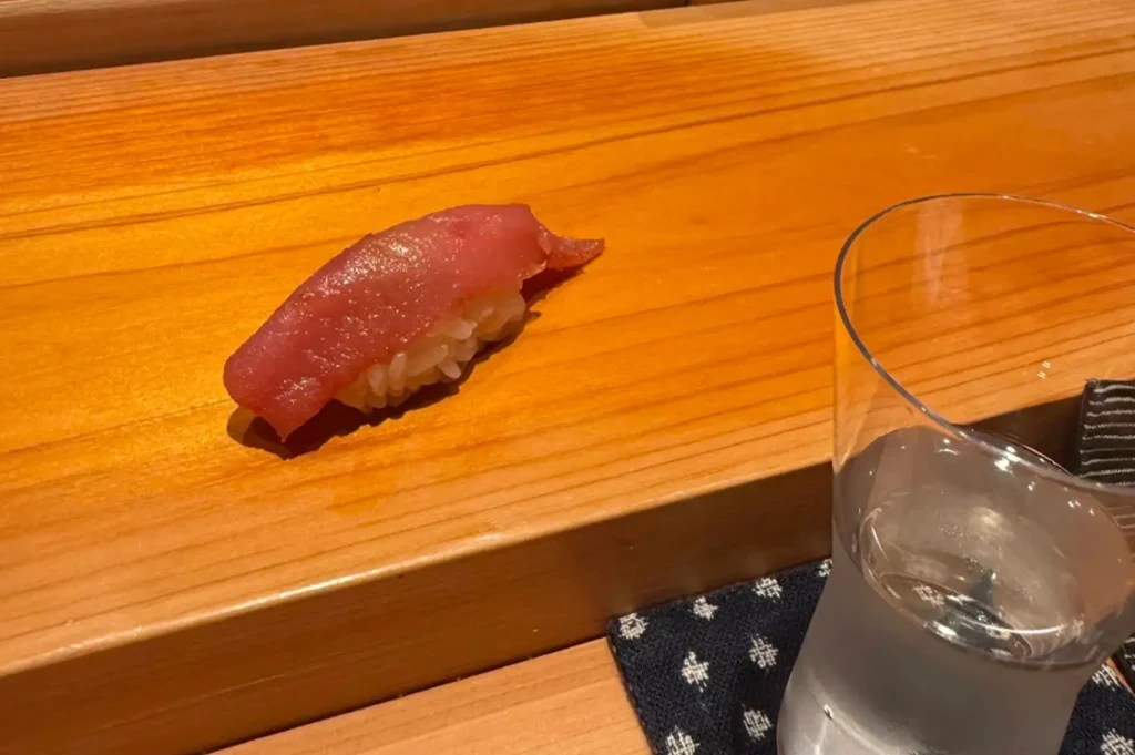 A high-angle shot showing a piece of tuna (maguro) nigiri placed on a wooden sushi board alongside a clear glass of water resting on a patterned blue cloth coaster. The piece features a glossy, deep-pink slice of raw tuna draped over tightly packed white sushi rice, casting a faint shadow on the warm wooden surface.