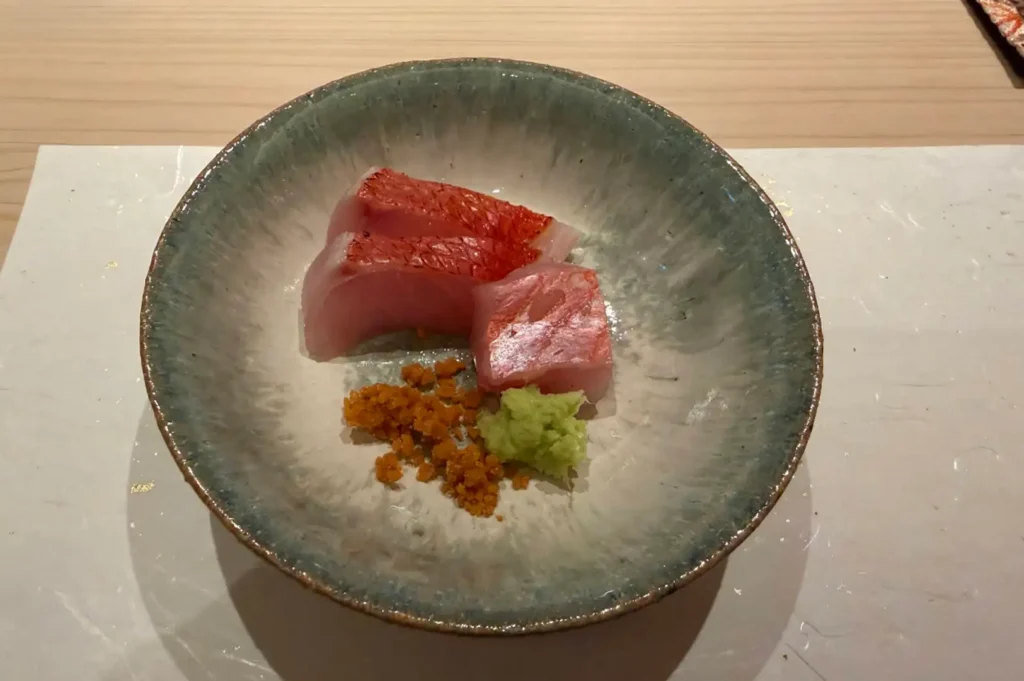 An overhead view presents three thick slices of red-skinned sashimi served in a rustic, speckled stoneware bowl over a clean white placemat. Small portions of freshly grated green wasabi and crunchy golden-brown toppings sit beside the fish, highlighting the vibrant color contrast and refined presentation.