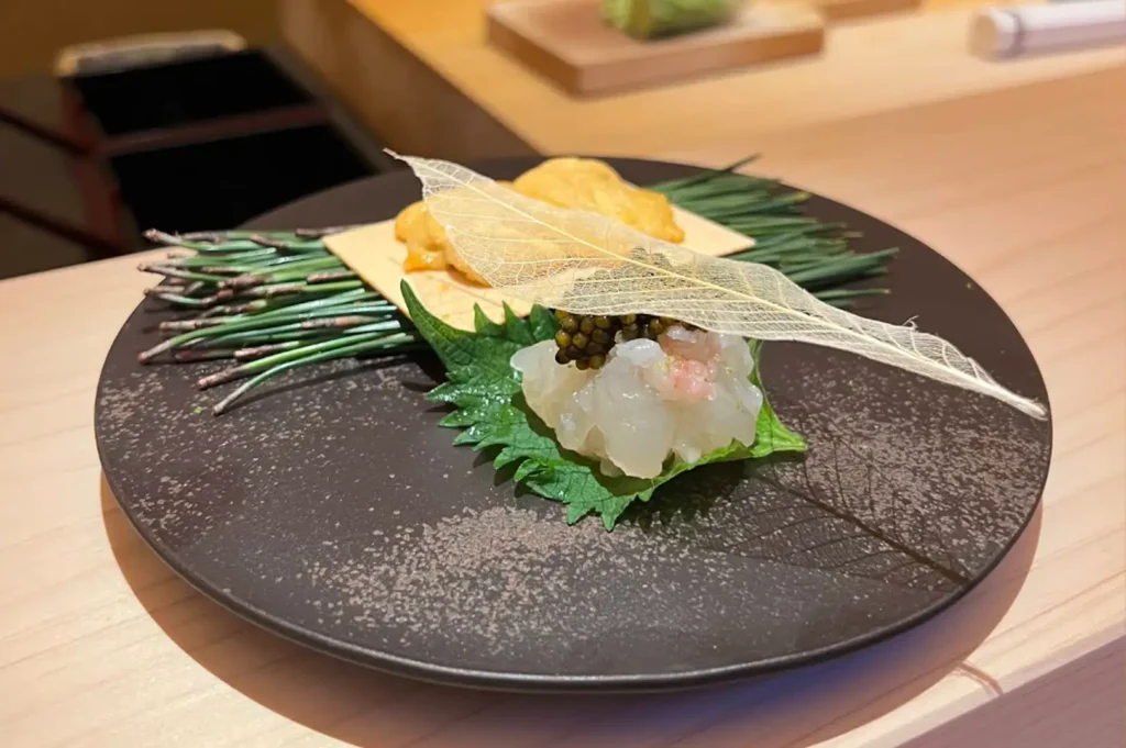 A elegant culinary presentation on a dark textured round ceramic plate resting on a wooden counter, featuring translucent raw white fish topped with caviar on a green shiso leaf, accompanied by bright yellow sea urchin (uni) on a crisp base, pine needles, and delicate translucent skeleton leaf garnishes.