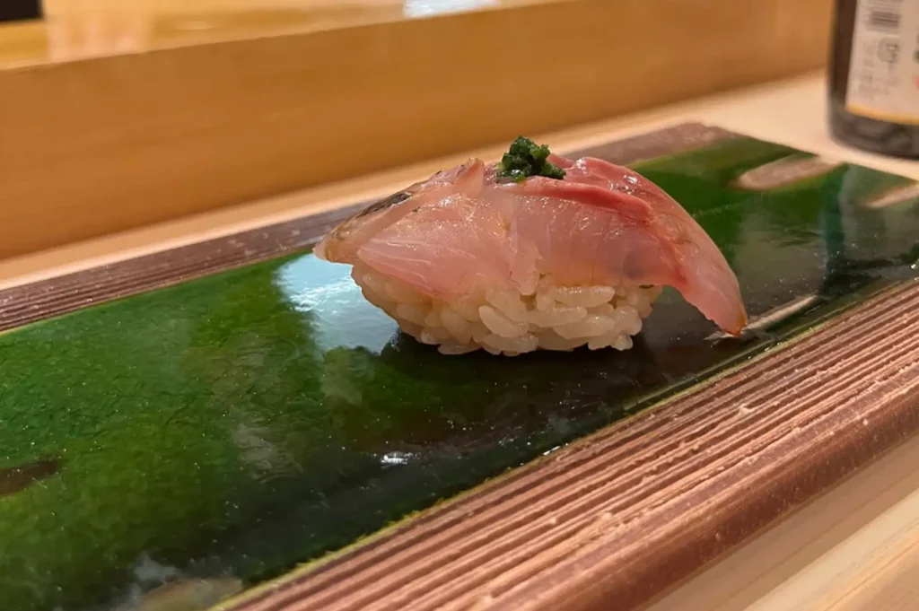 A close-up view of a piece of translucent, pinkish-white fish nigiri topped with fine green herbs, resting on seasoned sushi rice over a textured brown rectangular ceramic platter glazed with a glossy emerald-green center strip.