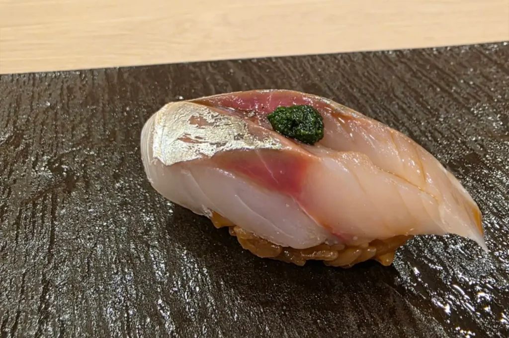 A piece of nigiri sushi features a slice of silver-skinned fish with a dab of dark green herb paste on top, resting over seasoned brown sushi rice on a textured, dark gray slate platter.