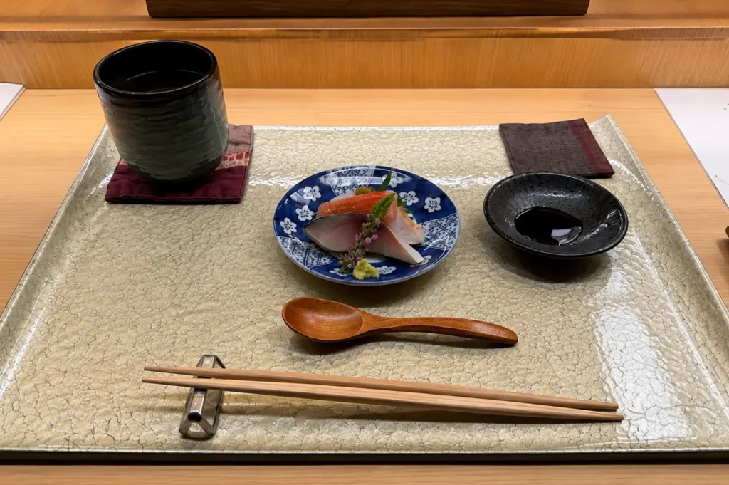 A minimalist place setting arranged on a crackle-glazed beige placemat features a blue-and-white floral ceramic dish holding sliced sashimi garnished with purple flower sprigs and fresh wasabi. To the sides sit a dark green tea cup on a fabric coaster, a small dish of soy sauce, a carved wooden spoon, and light wood chopsticks resting on a metallic rest.