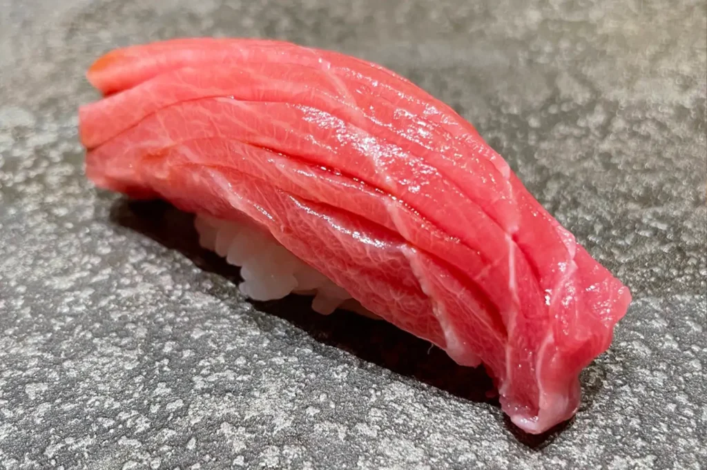 A macro photo highlights a single piece of otoro nigiri sushi resting on a dark gray, coarse stone platter. The thick slice of fatty tuna features prominent white marbling across its vibrant pink surface, expertly draped over a hand-molded bed of white sushi rice.