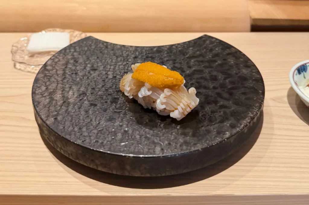 A single piece of intricate squid nigiri sushi topped with bright orange sea urchin (uni), served on a textured, crescent-shaped dark grey ceramic platter placed on a smooth wooden dining surface.