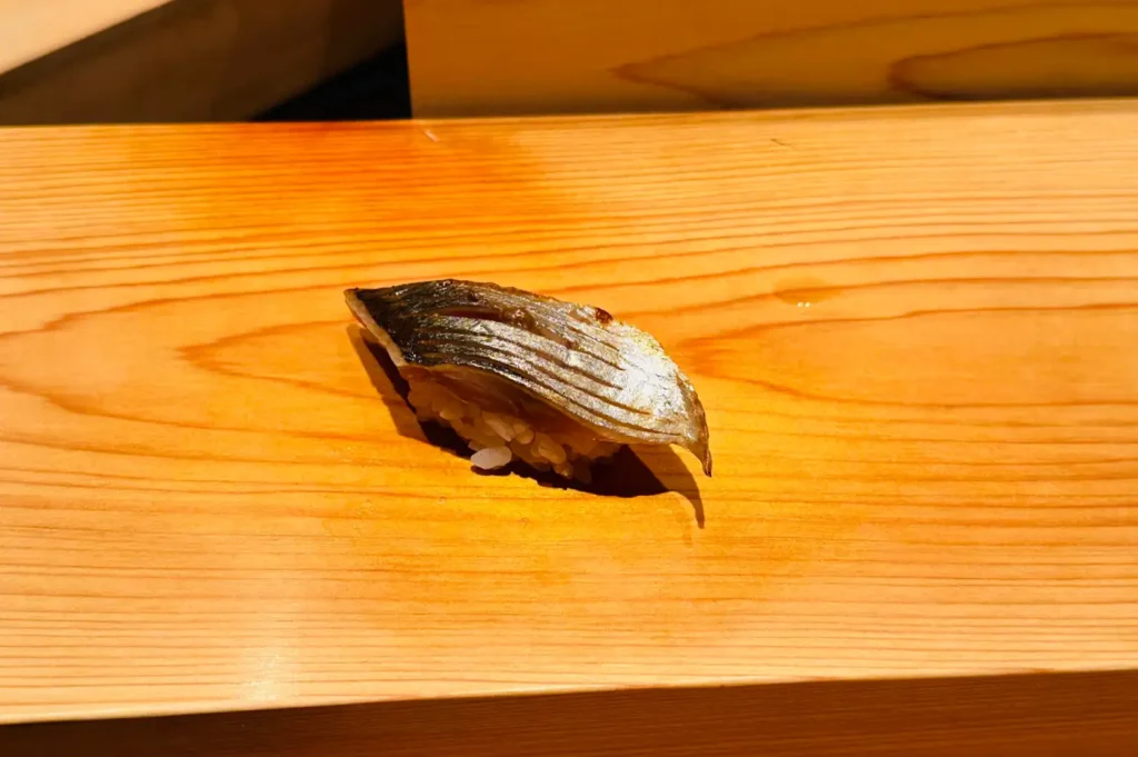 A close-up photograph of a single piece of seared mackerel (saba or kohada) nigiri resting on a light hinoki wood counter. The silvery skin of the fish features fine parallel score marks showing subtle charring from a blowtorch, gently curving over a bed of seasoned sushi rice.