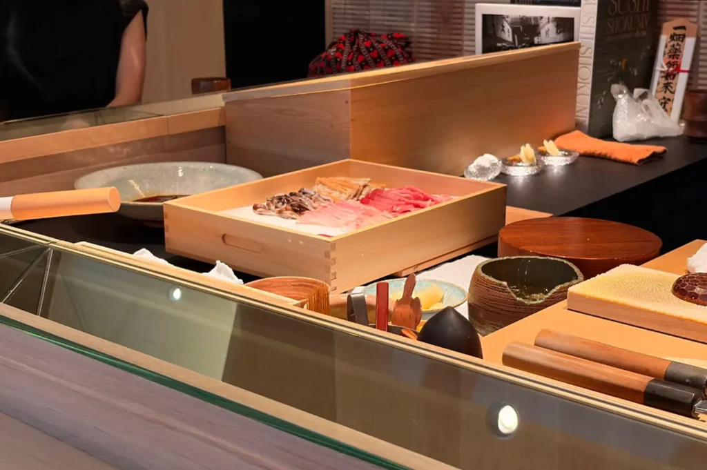 A perspective shot looking over the glass-topped sushi counter reveals the chef's workspace, complete with a light wooden tray holding neatly arranged slices of raw fish including tuna and white fish. Japanese culinary tools, including traditional knives with wooden handles, ceramic bowls, and condiment containers, line the counter top.