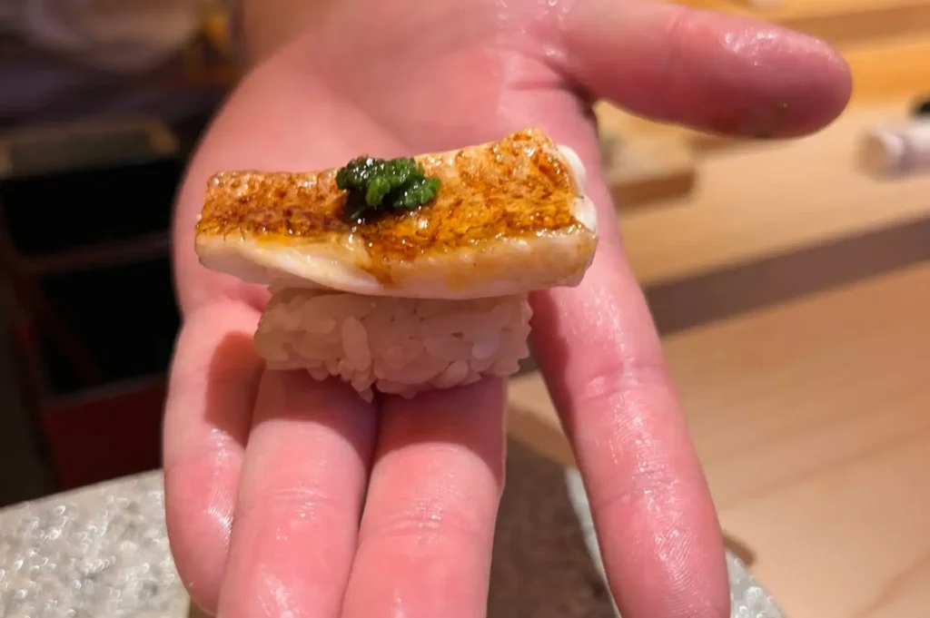 An intimate close-up shot capturing a sushi chef’s open palm offering a single piece of nigiri sushi, featuring a lightly charred, golden-brown fish piece resting over seasoned rice and topped with a small dab of vibrant green herb paste.
