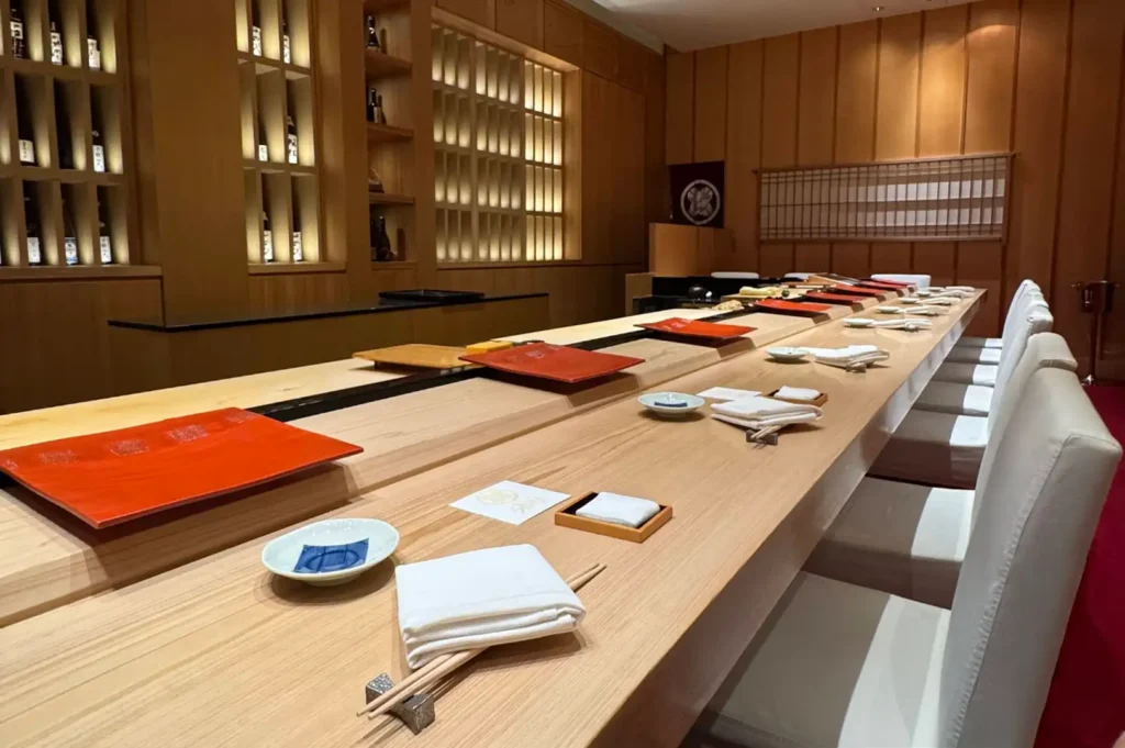 The interior of an omakase restaurant showcases a long, polished blonde-wood counter set with individual dining arrangements including white hand towels, wooden chopsticks, small sauce dishes, and bright orange lacquered serving boards. Modern off-white high-back chairs line the counter, backed by warm ambient lighting, wooden screen walls, and built-in shelving displaying bottles of sake.