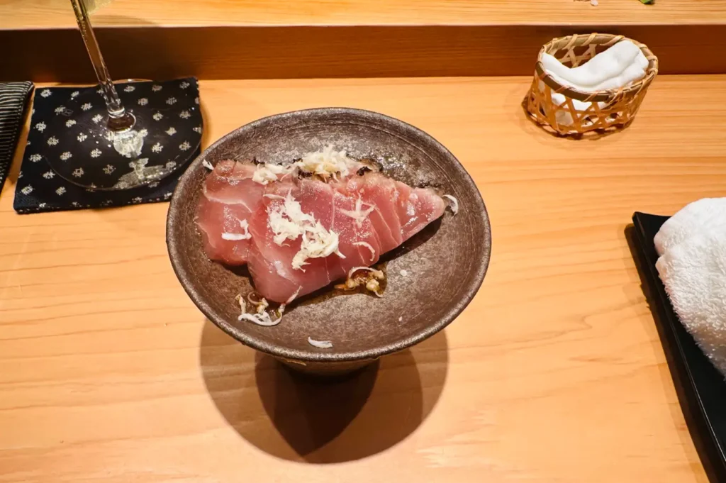 A top-down shot of a dark, rustic ceramic pedestal bowl containing slices of raw, lightly seared bonito or tuna sashimi drizzled with a light sauce and topped with finely shredded white radish or ginger. The dish sits on a smooth wooden table next to a patterned dark coaster holding the base of a wine glass, a woven basket with a rolled cloth, and a folded white hand towel.