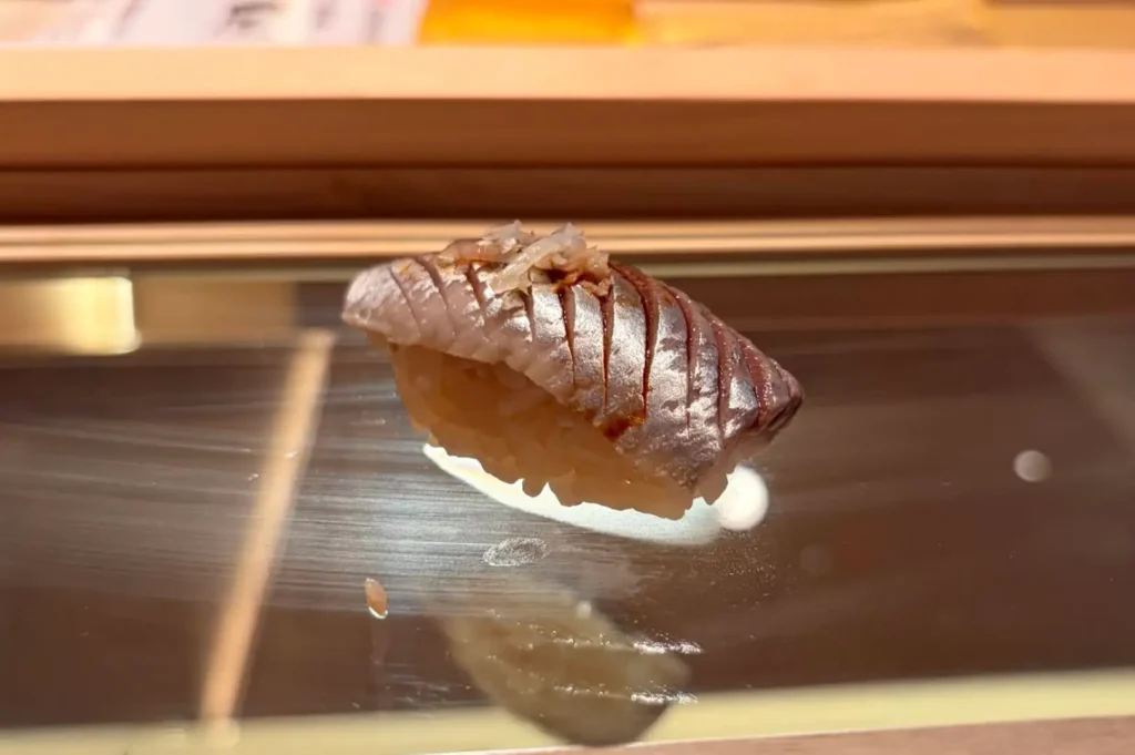 An extreme close-up shot focuses on a piece of nigiri sushi placed on a sleek, dark glass display ledge in a Japanese omakase restaurant. The sushi features a slice of silver-skinned fish scored with precise parallel diagonal incisions, brushed with soy glaze, topped with delicate shredded garnish, and resting on a hand-molded mound of seasoned rice.