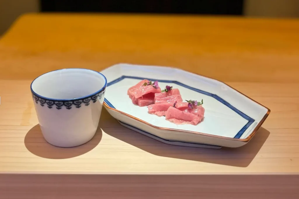 A eye-level table arrangement set on a smooth light-wood surface, displaying the hexagonal blue-rimmed ceramic dish with three pieces of flower-garnished pink tuna sashimi alongside a matching white porcelain tea or sake cup detailed with a blue decorative rim pattern.