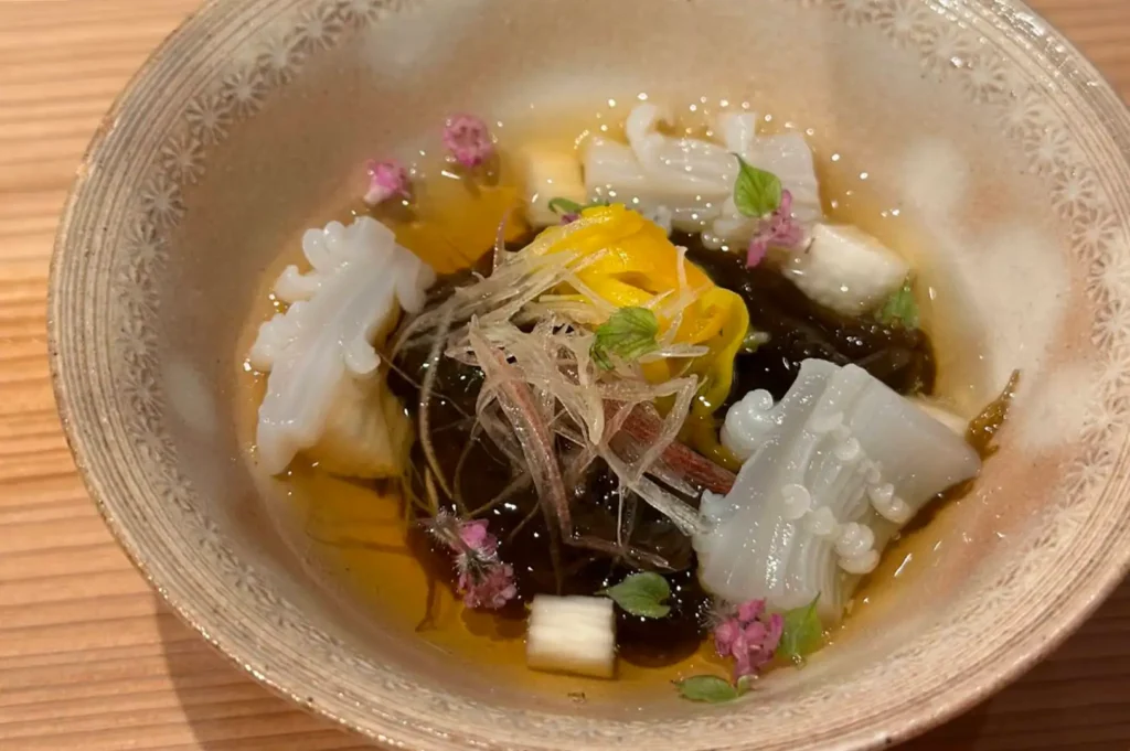 A shallow stoneware bowl filled with a delicate chilled broth containing tender pieces of scored squid, mozuku seaweed, shredded ginger, bright yellow flower petals, and tiny pink edible blossoms.