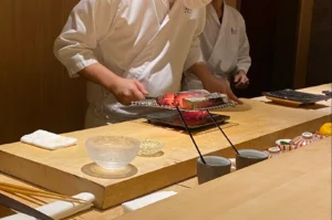 A chef dressed in a white chef's coat and protective face mask stands behind a light wooden sushi counter, using a metal mesh grill held over long chopsticks to sear a piece of fish, with glass bowls and ceramic condiment vessels neatly arranged nearby on the counter.