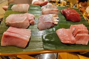 A high-angle close-up of a wooden platter lined with fresh green banana leaves, showcasing an array of meticulously sliced raw fish for omakase preparation. The spread includes various premium cuts of seafood, ranging from delicate white fish and silver-skinned slices to vibrant ruby-red tuna and marbled pink otoro.