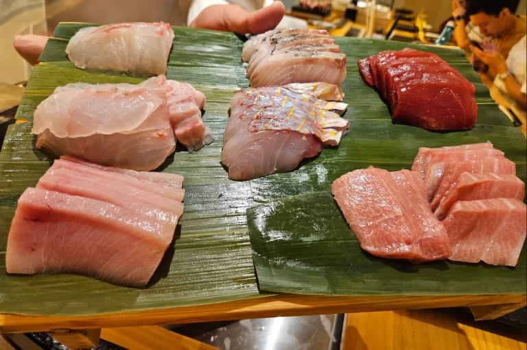 A high-angle close-up of a wooden platter lined with fresh green banana leaves, showcasing an array of meticulously sliced raw fish for omakase preparation. The spread includes various premium cuts of seafood, ranging from delicate white fish and silver-skinned slices to vibrant ruby-red tuna and marbled pink otoro.