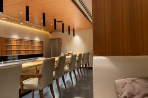 A wide view of a modern Japanese omakase dining room featuring a sleek light-wood counter lined with padded high-backed chairs, illuminated by sleek black pendant lights hanging from a warm wooden ceiling panel. The surrounding minimalist space is decorated with neutral wall textures, traditional wooden lattice screen panels, and plush light-beige banquette seating in the foreground.