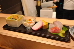 A long rectangular black plate presented on a wooden sushi counter displays a row of omakase appetizers, including a square ceramic cup filled with greens and bonito flakes, a small glass dish holding delicate tofu topped with salmon roe, a slice of pink-rimmed kamaboko, a piece of tamagoyaki, fresh sashimi cuts arranged over a green leaf, and a mound of fresh wasabi.