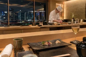 An intimate viewpoint from a diner's seat at a light wood omakase counter overlooking a panoramic night view of the illuminated city skyline through floor-to-ceiling windows. In the foreground, a black textured stoneware plate holds two pieces of delicate nigiri topped with edible purple flowers and a small mound of pickled ginger, flanked by a green ceramic tea cup, a glass of white wine, a rolled hand towel, and a covered soup bowl. In the background, a sushi chef wearing a white traditional uniform stands focused at his preparation station under warm ambient lighting.
