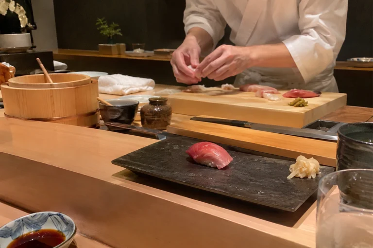 A close-up view from a diner’s perspective at an omakase counter, featuring a freshly prepared piece of tuna nigiri on a dark slate plate alongside pickled ginger. In the background, a sushi chef wearing a white jacket meticulously shapes a piece of sushi on a thick wooden prep board arranged with raw fish cuts, a wooden rice tub, and traditional ceramic soy pots.