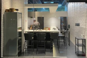 A view from outside a glass storefront reveals the sleek interior of Ryo Sushi in Tanjong Pagar, featuring an L-shaped white dining counter lined with modern high-backed dark gray stools. Inside, a chef works behind the counter in a kitchen area enclosed by white subway-tiled walls, while a tall glass-door drink refrigerator stands near the front entrance.