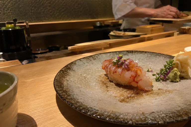 A close-up, eye-level diner’s view showcases a piece of botan ebi nigiri sushi delicately art-drizzled and garnished with small purple flowers, served on a wide, textured ceramic plate. Next to the piece sits a mound of wasabi, shiso blossoms, and pickled ginger, set on a warm wooden omakase counter while a chef in a white chef coat prepares the next course in the softly lit, blurred background.