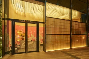The entrance of Sushi Masaaki features double glass doors with dark frames, topped with bold lettering spelling out the restaurant's name. Warm light illuminates the interior, revealing a marble reception counter, vibrant pink wallpaper patterned with white cranes, and a striking suspended wooden ceiling sculpture made of undulating wave-like slats that extend across the glass facade behind wooden vertical blinds.